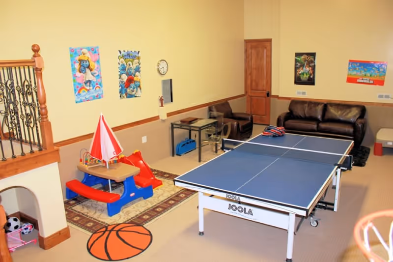 A recreational room with a blue JOOLA ping pong table in the center, a brown leather couch and chair against the far wall, children's play area with a small red and blue picnic table and slide, colorful posters on the beige walls, and a basketball-themed rug on the carpeted floor.