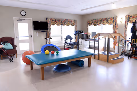 A bright therapy or exercise room with a padded table in the center holding three small colored balls. Around the room are various exercise and therapy equipment including a large red exercise ball, a blue trampoline, a balance trainer, parallel bars, and a walker. The room has windows with patterned valances, a wall clock, and a door with a window.