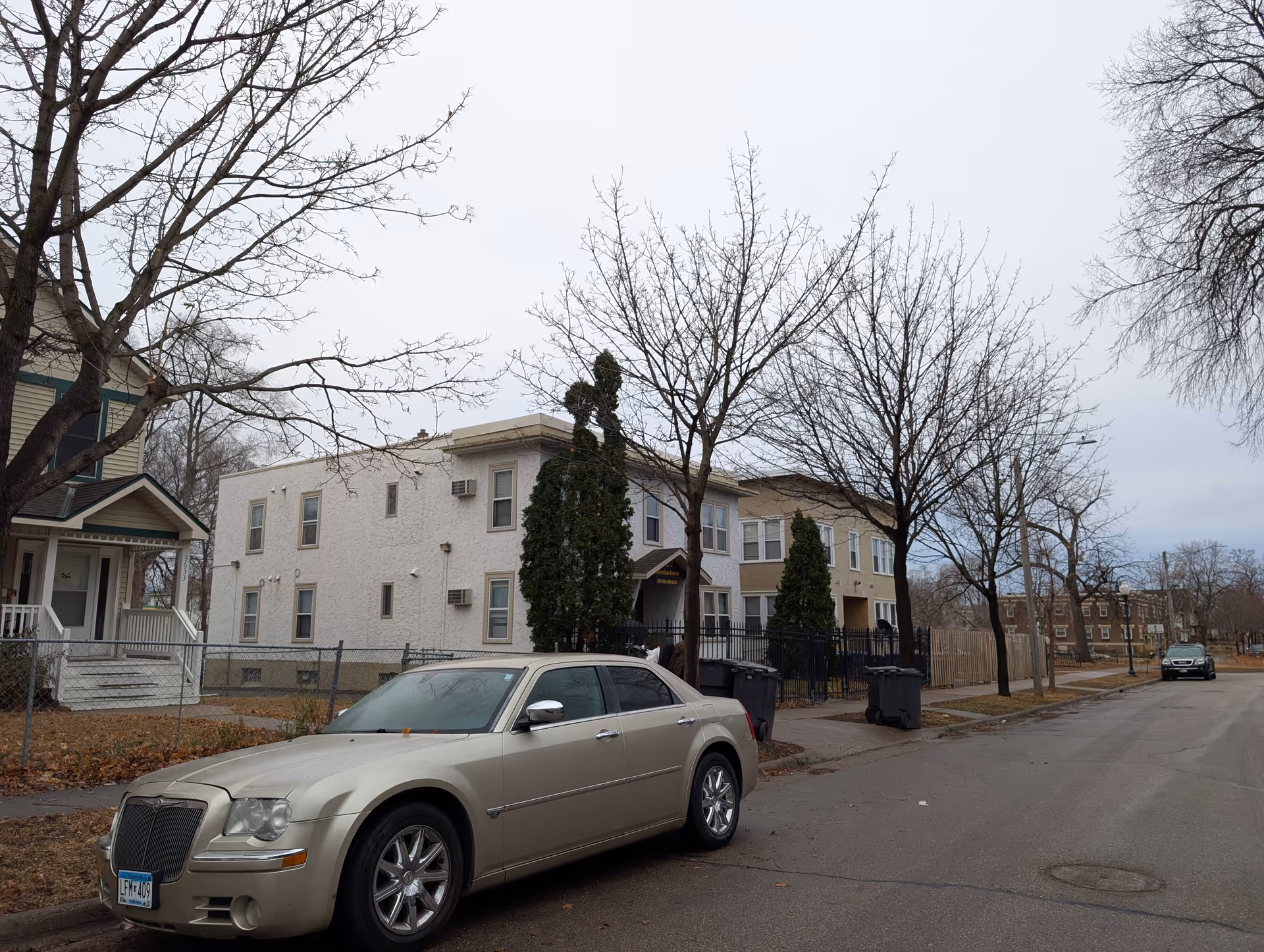 Street view showing a beige sedan parked on the side of a residential street with leafless trees and multi-unit houses in the background under an overcast sky.
