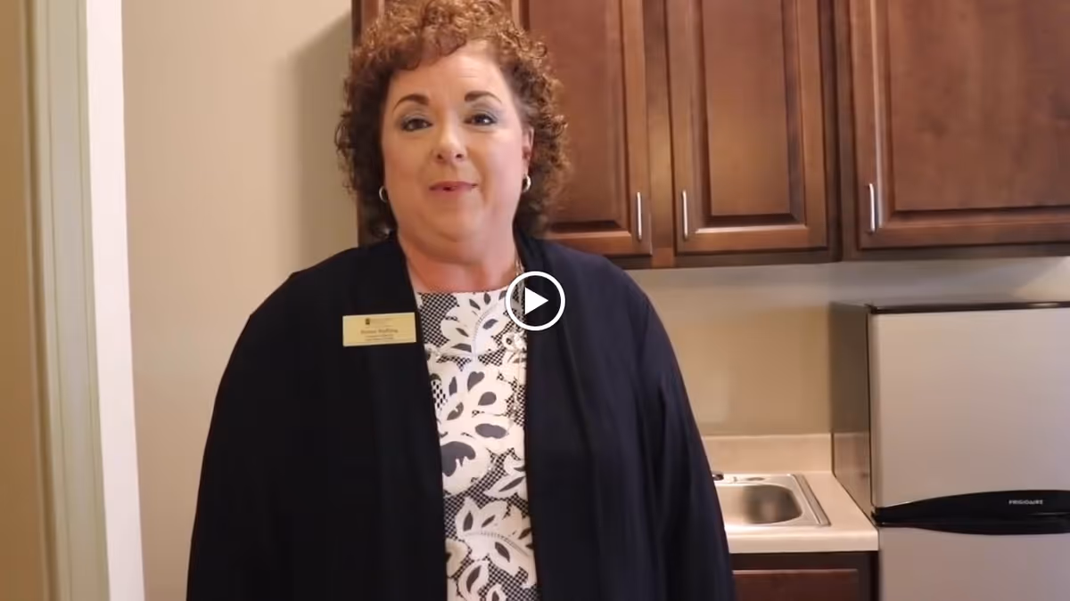 A woman with curly hair wearing a black cardigan and a patterned blouse stands in a kitchen area with wooden cabinets, a small sink, and a compact refrigerator behind her.