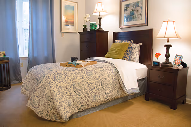 A cozy bedroom with a single bed covered in a patterned beige and blue bedspread. The bed has multiple pillows, including a green one. There is a wooden nightstand with a lamp, a small flower vase, a digital clock, and a framed photo. A wooden dresser with a lamp, a small plant, and decorative items is against the wall. The room has beige carpet, light-colored walls, two framed pictures, and a window with blue curtains.
