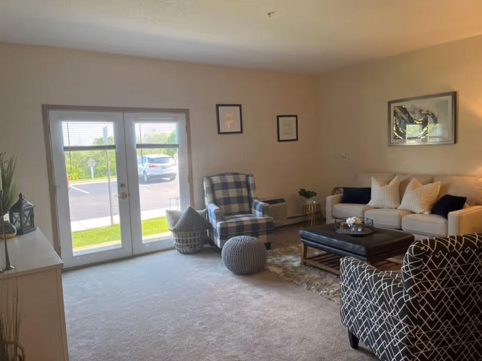 A cozy living room with a beige carpet, a white sofa with multiple pillows, two patterned armchairs, a large ottoman coffee table, and a small round side table with a plant. French doors with blinds lead outside to a parking lot with a car visible. The walls are decorated with framed artwork.