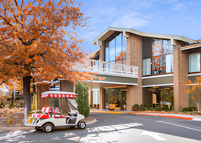 Exterior view of a senior living facility building with large windows and a covered entrance. A golf cart with a red and white striped canopy is parked near a tree with orange autumn leaves.