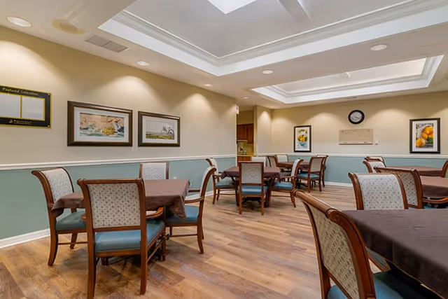 A dining room with several tables covered with brown tablecloths and surrounded by wooden chairs with light patterned upholstery and blue cushions. The room has light green and beige walls, framed artwork, a clock, and a skylight ceiling providing natural light. The floor is wooden.