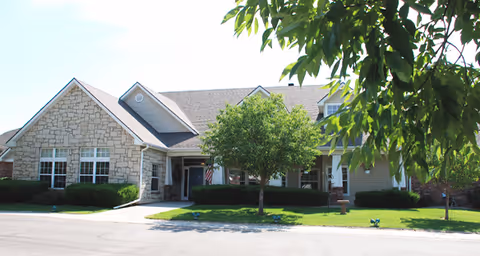 Exterior view of a single-story building with a stone and siding facade, surrounded by green grass and trees under a clear sky.