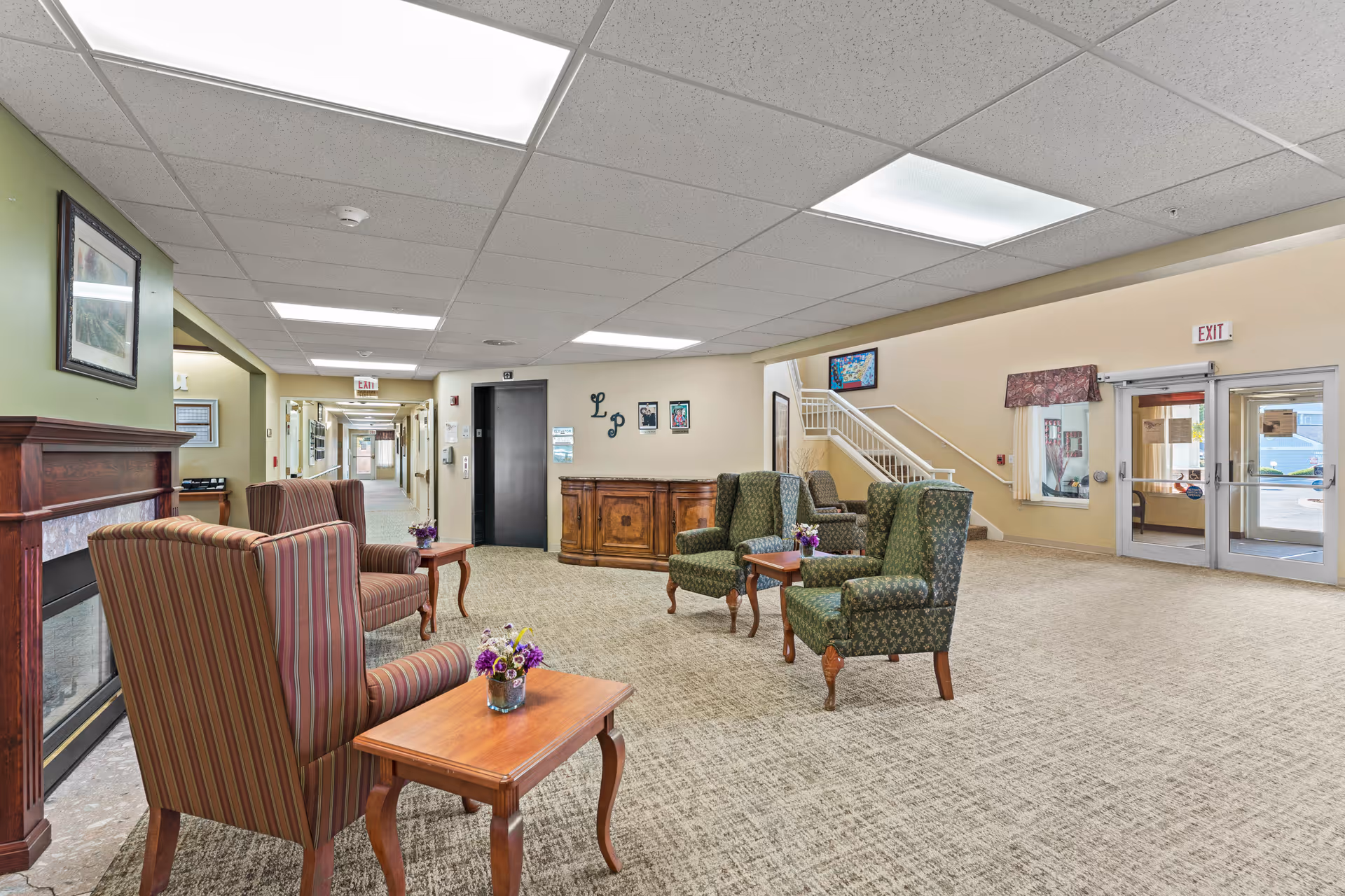 A spacious assisted living facility common area with several upholstered armchairs and wooden side tables with small flower arrangements. There is a fireplace on the left wall, a hallway leading to rooms in the background, an elevator, and a staircase near the entrance doors on the right. The room is well-lit with ceiling lights and has carpeted flooring.