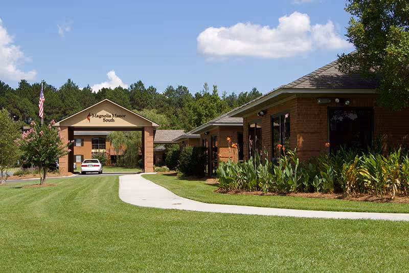 Exterior view of Magnolia Manor South facility showing a brick building with large windows, a covered entrance with the facility name, a white car parked near the entrance, a well-maintained lawn, a concrete walkway, and trees in the background under a partly cloudy sky.