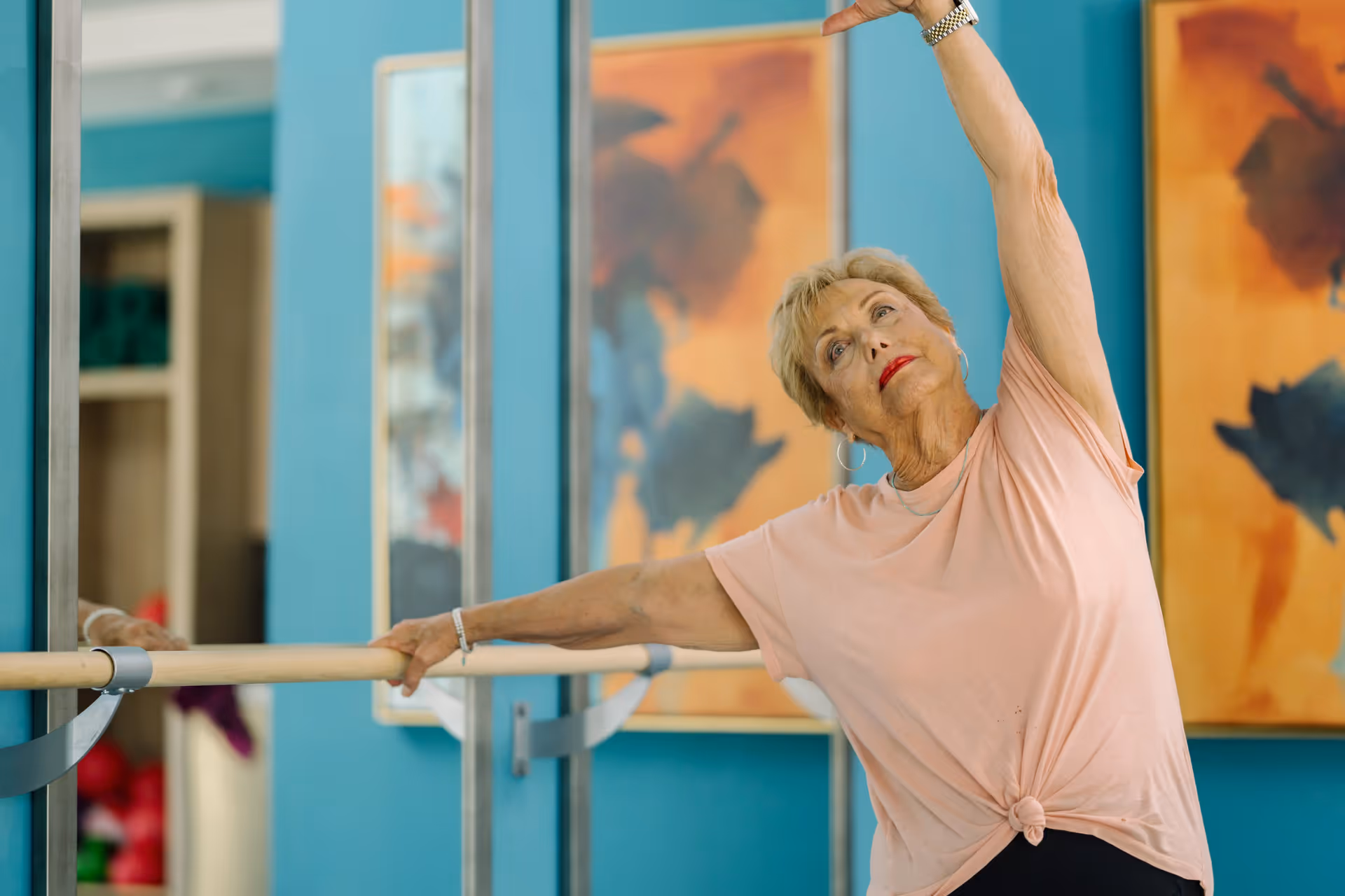 An elderly woman in a light pink shirt stretching her arm while holding onto a ballet barre in a room with blue walls and abstract orange and black paintings.