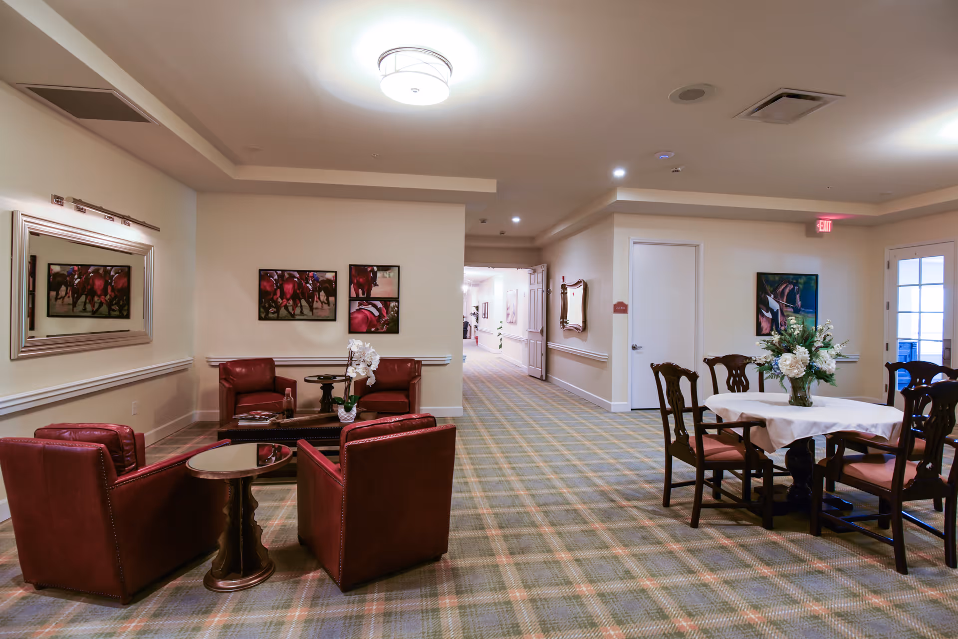 A cozy common area in a senior living facility with four red leather armchairs arranged around a small round table on the left, and a round dining table with a white tablecloth and floral centerpiece surrounded by six wooden chairs on the right. The room has beige walls, a plaid carpet, framed artwork, and a large mirror on the left wall. A hallway extends in the background with additional artwork and lighting.