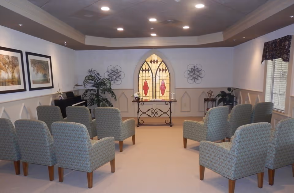 A small chapel or meditation room with two rows of upholstered chairs facing a decorative stained glass window. The room has beige walls with wainscoting, two framed landscape paintings on the left wall, a piano in the back left corner, and a window with a valance on the right. There are also some plants and decorative metal wall art.