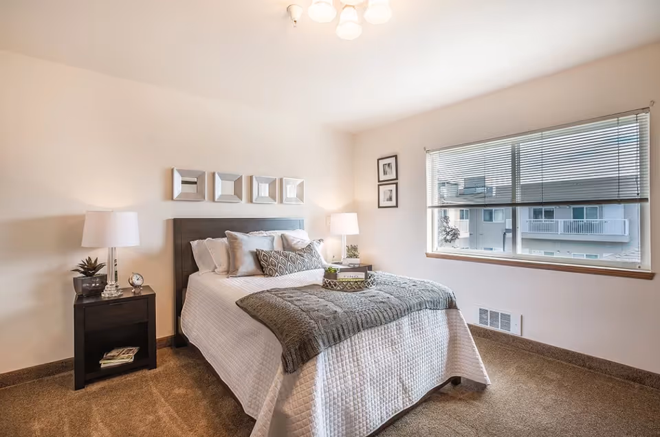 A neatly made bed with white bedding and a gray throw blanket in a bedroom. There are two bedside tables with lamps and decorative items, a large window with blinds, and framed pictures on the walls.