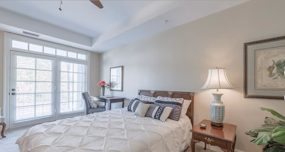 A bright bedroom with a large bed covered in a white textured comforter and multiple pillows. Next to the bed is a wooden nightstand with a decorative lamp and a small item. A framed picture hangs on the wall above the nightstand. On the opposite side, there is a small wooden desk with a chair, a vase with pink flowers, and a mirror above it. Large glass doors with white blinds allow natural light to fill the room.