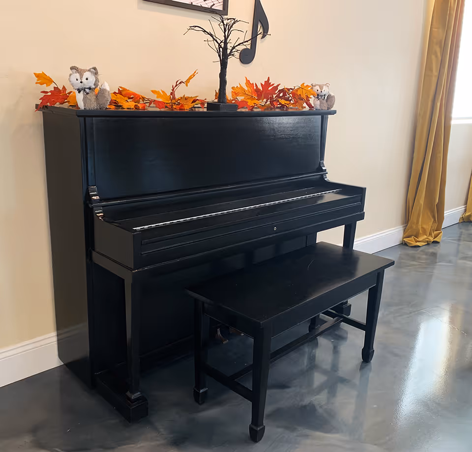A black upright piano with a matching bench in front of it, decorated with autumn leaves and two small stuffed animal figures on top. A decorative black tree and a musical note wall decoration are visible above the piano. The room has a polished concrete floor and beige walls with a window covered by mustard yellow curtains.