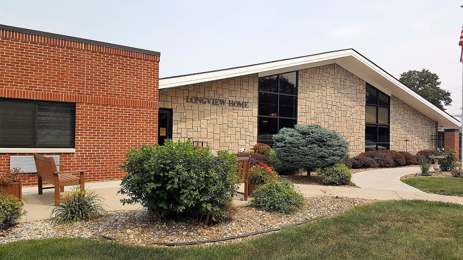 Exterior view of a senior living facility named Longview Home, featuring a building with a combination of red brick and stone walls, large windows, a landscaped garden with bushes and flowers, a concrete walkway, and wooden benches.