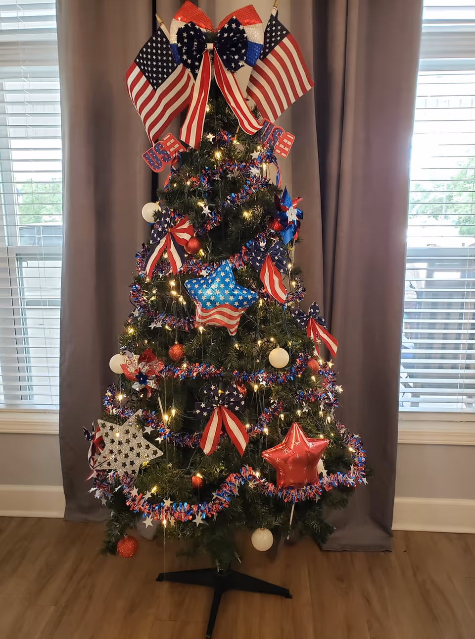 A patriotic, American-flag themed Christmas tree with star and bow ornaments and lights in front of curtains and windows.