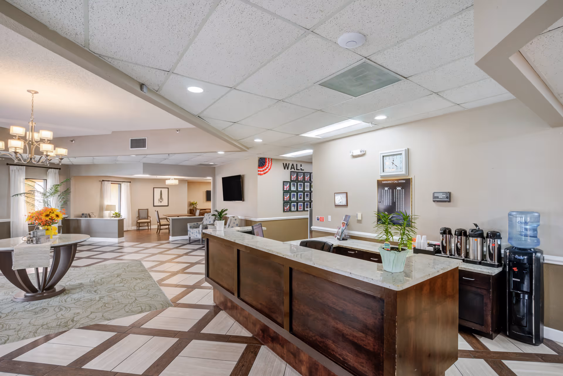 Reception area of a senior living facility with a wooden front desk, a water dispenser, coffee station, and a clock on the wall. The space has patterned tile flooring, a chandelier over a round table with a flower arrangement, and seating areas in the background.
