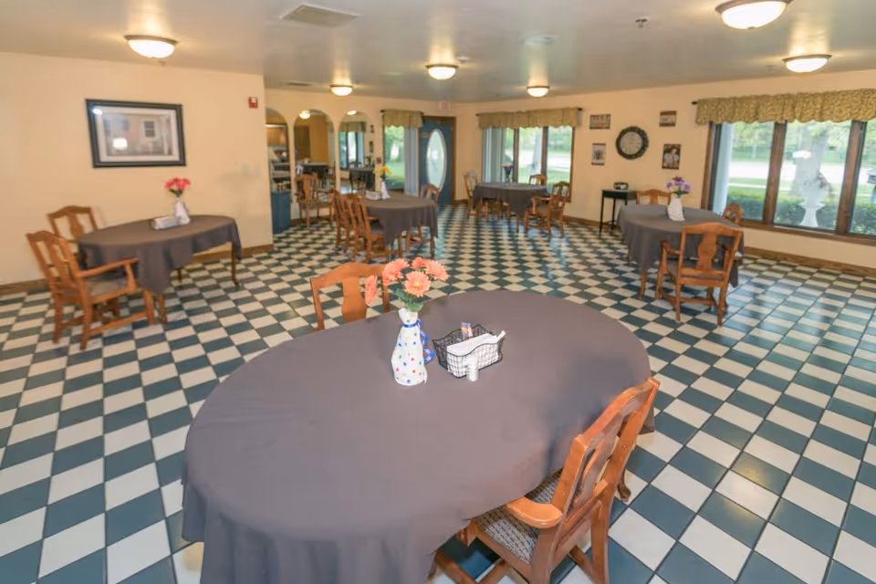 A dining room with several wooden tables covered with dark tablecloths and wooden chairs around them. Each table has a vase with flowers. The floor has a checkered pattern with green and white tiles. Large windows on one side let in natural light, and there are framed pictures and a clock on the walls.