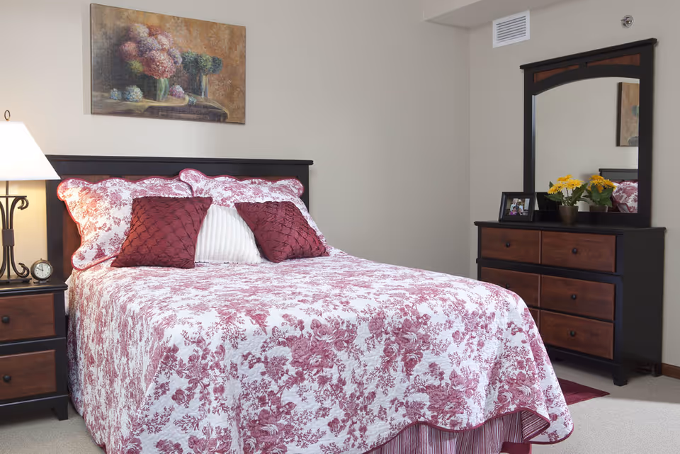 A neatly made bedroom featuring a bed with a red-and-white floral quilt, a nightstand with a lamp, and a dresser with mirror and flowers.