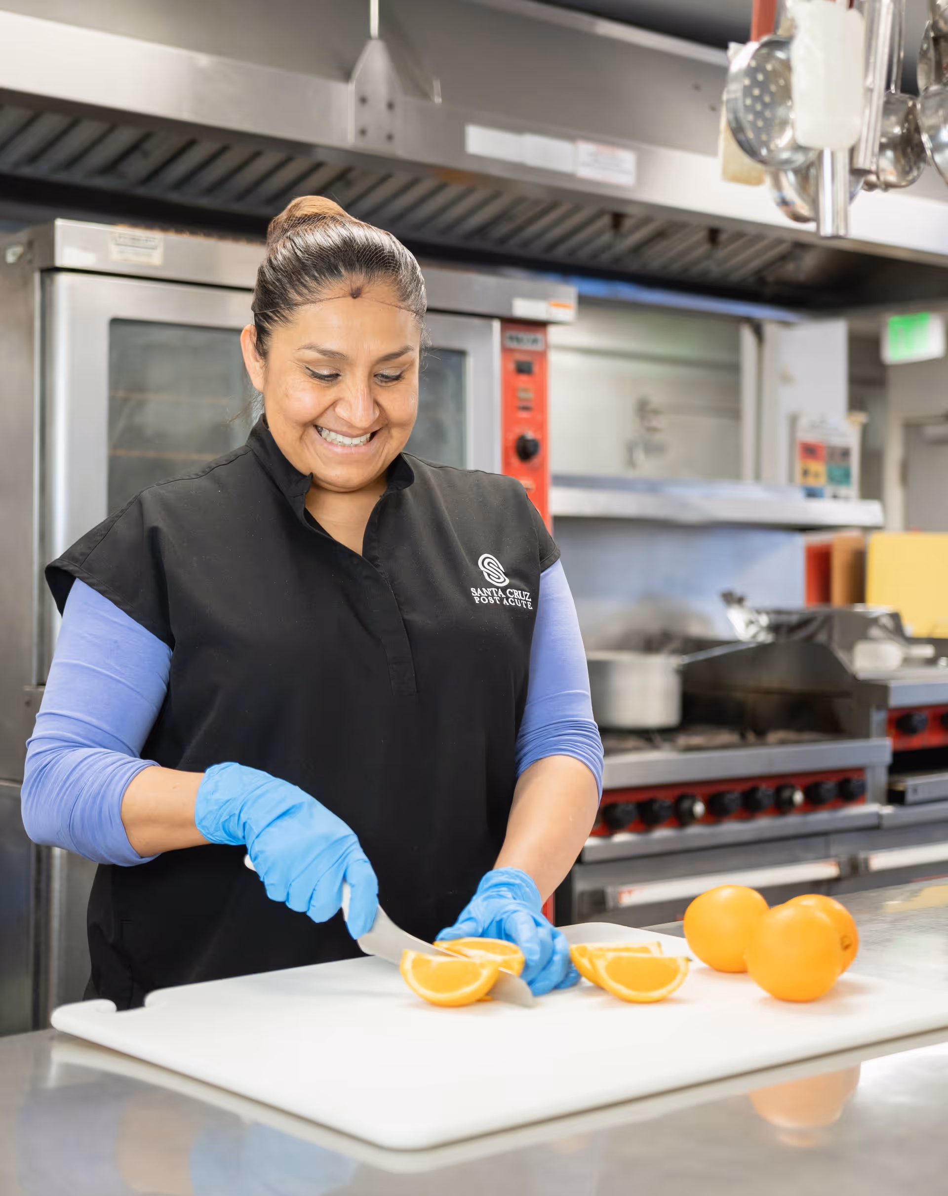 A woman wearing blue gloves and a black vest with the Santa Cruz Post Acute logo is smiling while slicing an orange on a white cutting board in a commercial kitchen.