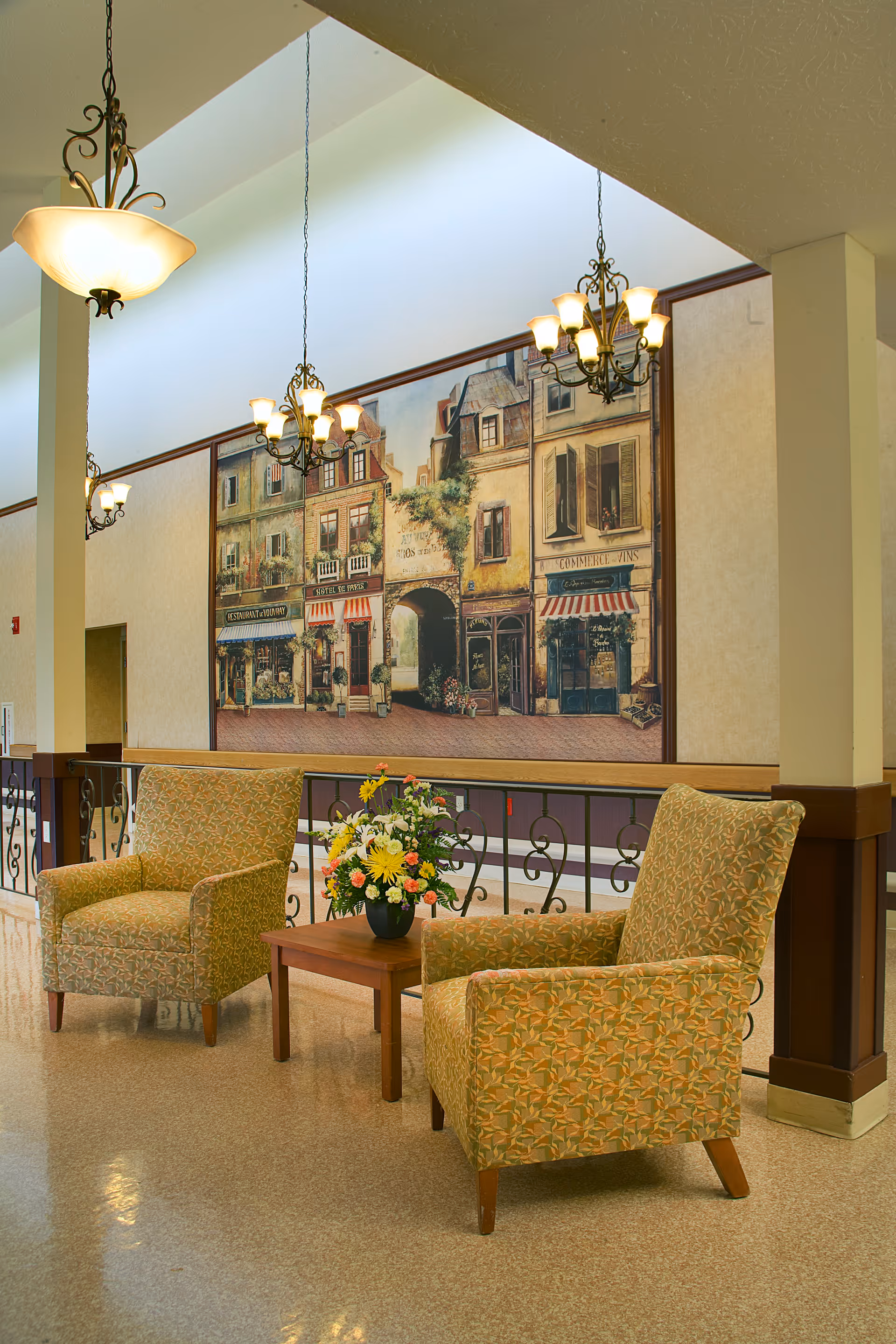 A cozy seating area with two patterned armchairs and a small wooden table with a flower arrangement in a senior living facility. The background features a large mural depicting a quaint European street scene with buildings and shops. Elegant hanging light fixtures illuminate the space.