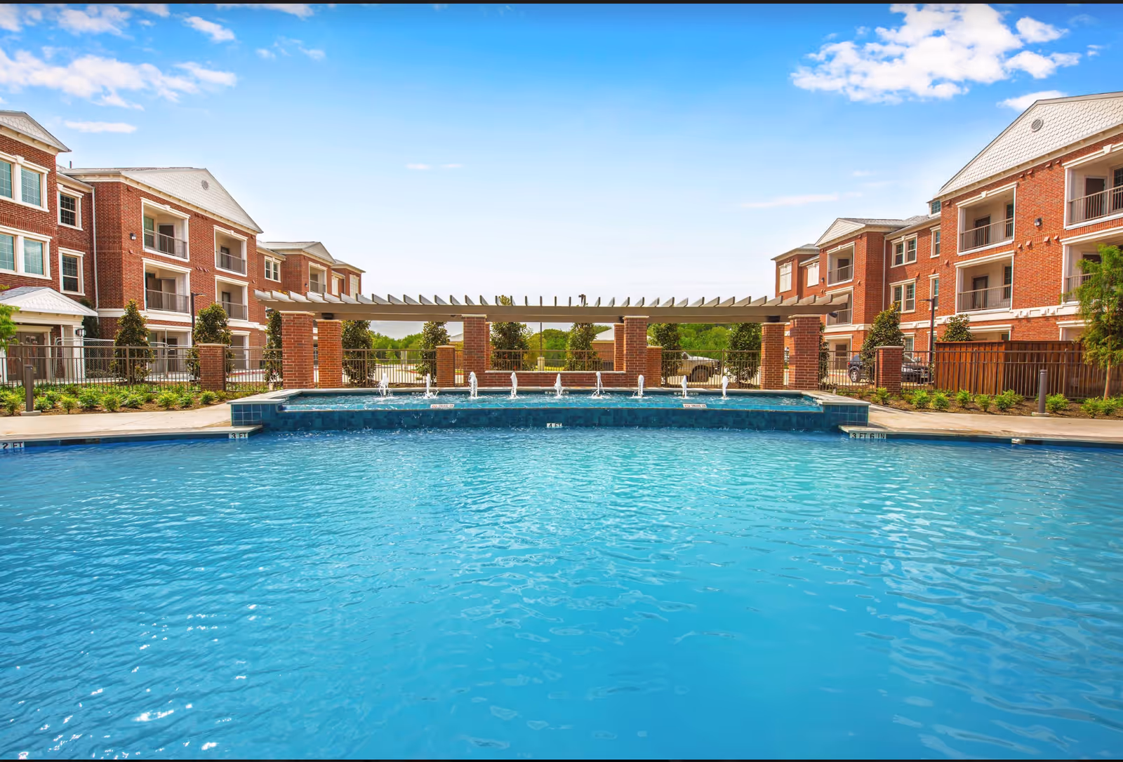 Outdoor swimming pool with clear blue water in front of a multi-story brick residential building under a partly cloudy sky. The pool area features a pergola with water fountains along the edge and landscaped greenery around the perimeter.