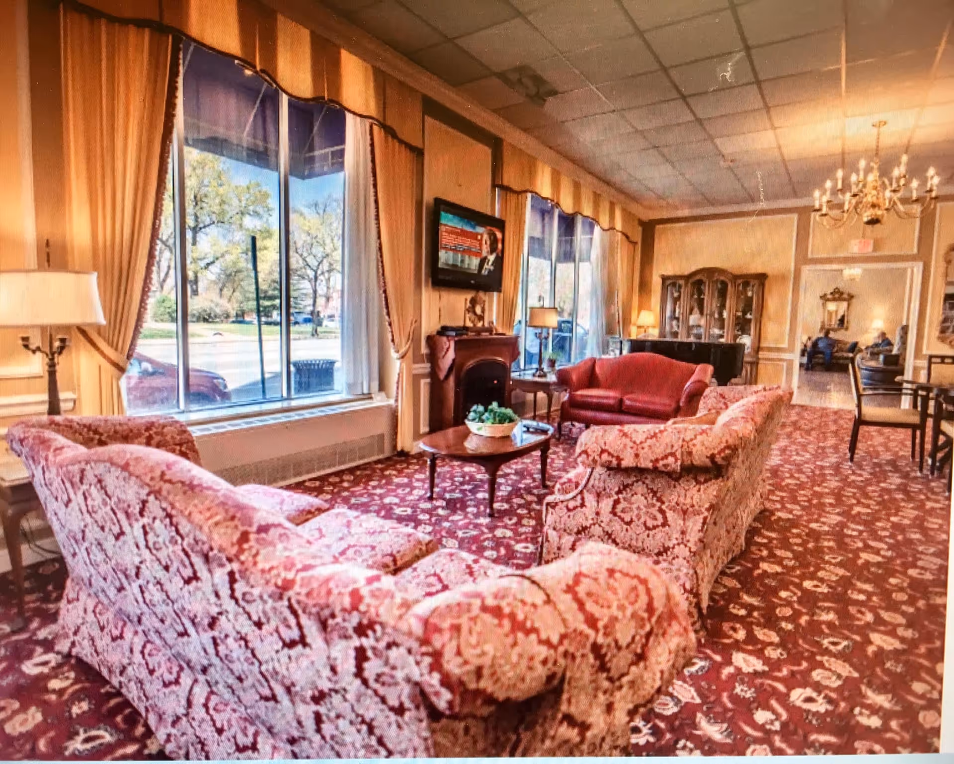 A spacious, elegantly decorated living room with patterned red and beige sofas arranged around a wooden coffee table. Large windows with yellow curtains allow natural light to fill the room. The room features a red carpet with floral patterns, a chandelier, a wall-mounted TV, and a wooden cabinet in the background.