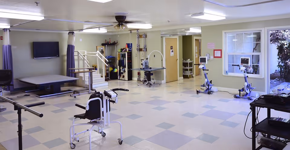 Spacious rehabilitation therapy room with exercise and mobility equipment, parallel bars, stationary bikes, a treatment table, and a wall-mounted TV.