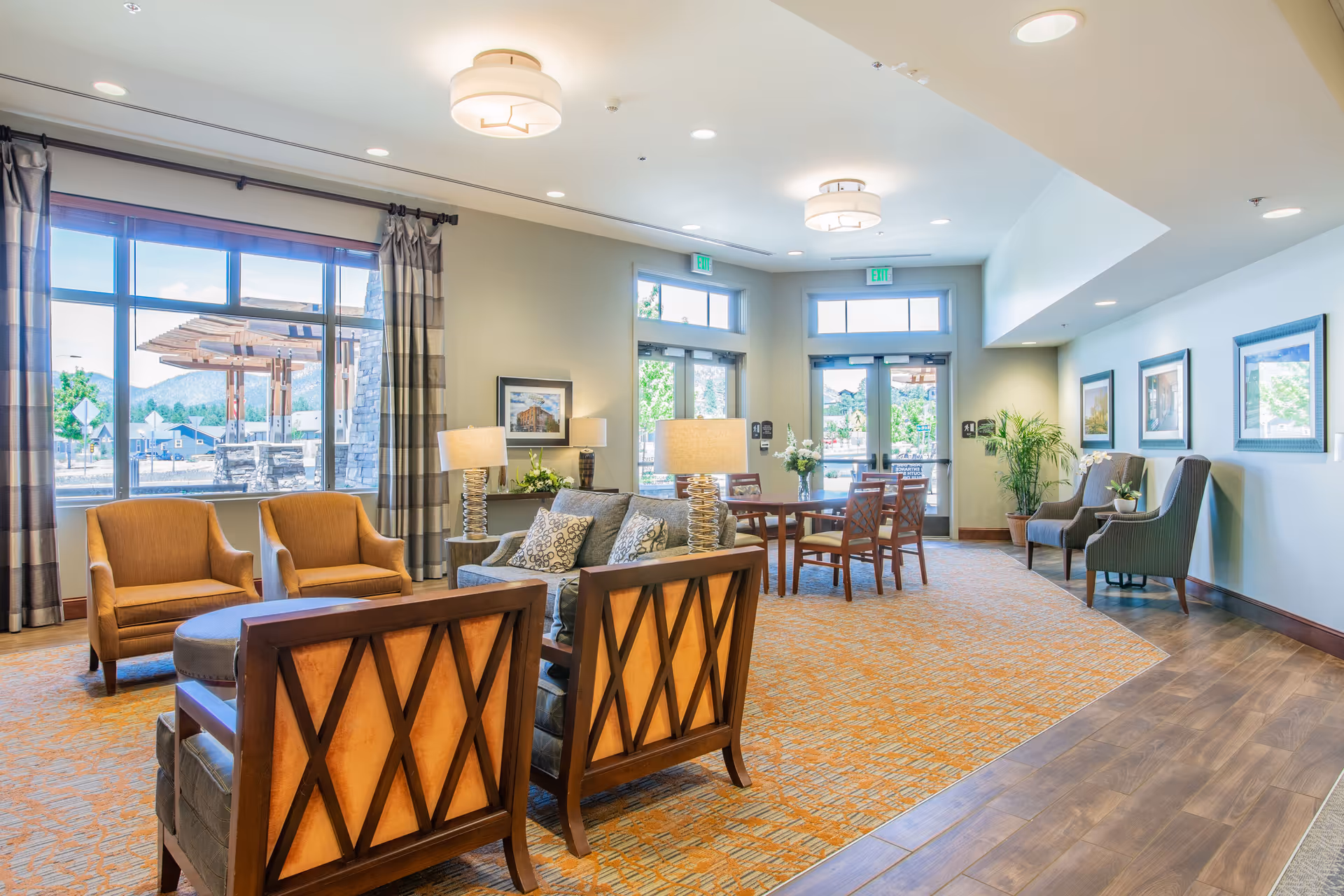 Bright, spacious lounge area with upholstered chairs, sofas, tables, lamps, and large windows by the entry doors.