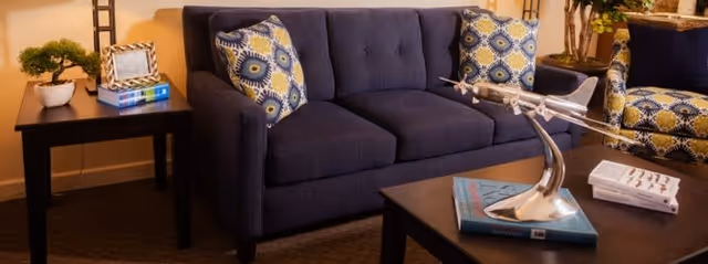 A cozy living room area with a dark blue sofa adorned with patterned cushions, a matching armchair, a dark wooden coffee table with a decorative silver airplane sculpture and books, and a side table with a small potted plant and picture frame.