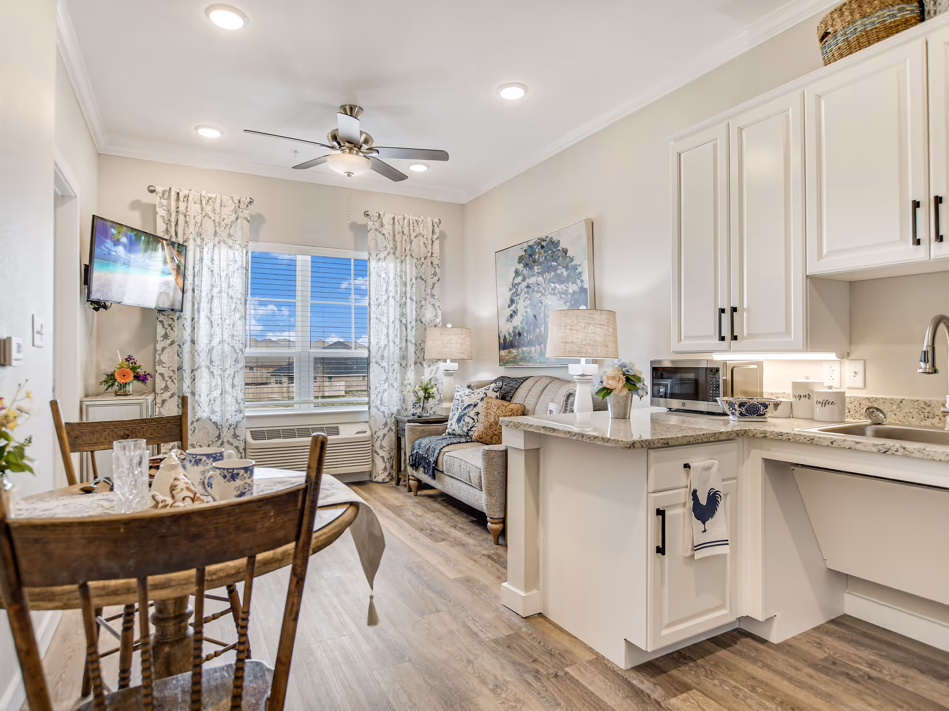 A bright and cozy living space featuring a small kitchen with white cabinets and granite countertops, a dining table with wooden chairs, a sofa with decorative pillows, two table lamps, a wall-mounted TV, and a large window with patterned curtains letting in natural light.