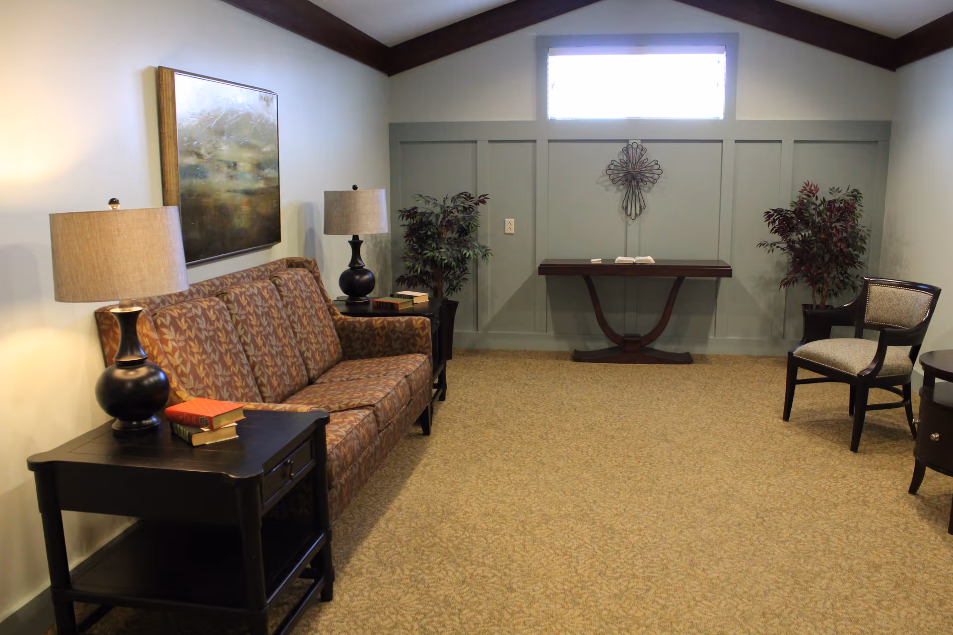 A cozy living room area with a patterned brown sofa, two black side tables each with a lamp and books, a decorative painting on the wall above the sofa, a wooden console table against a paneled wall with a metal wall decoration above it, two potted plants, and a single upholstered chair on the right side.