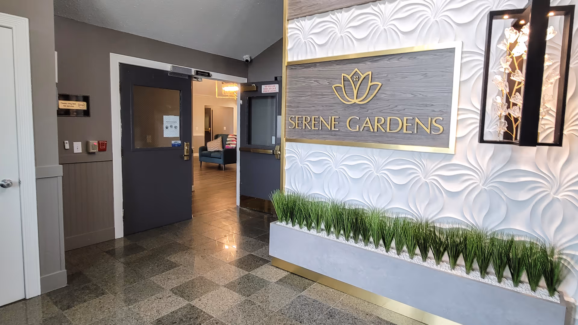 Entrance area of a senior living facility with a decorative wall featuring a sign that reads 'Serene Gardens' above a planter with green plants. The floor is tiled, and there are double doors leading to a lounge area with seating visible in the background.