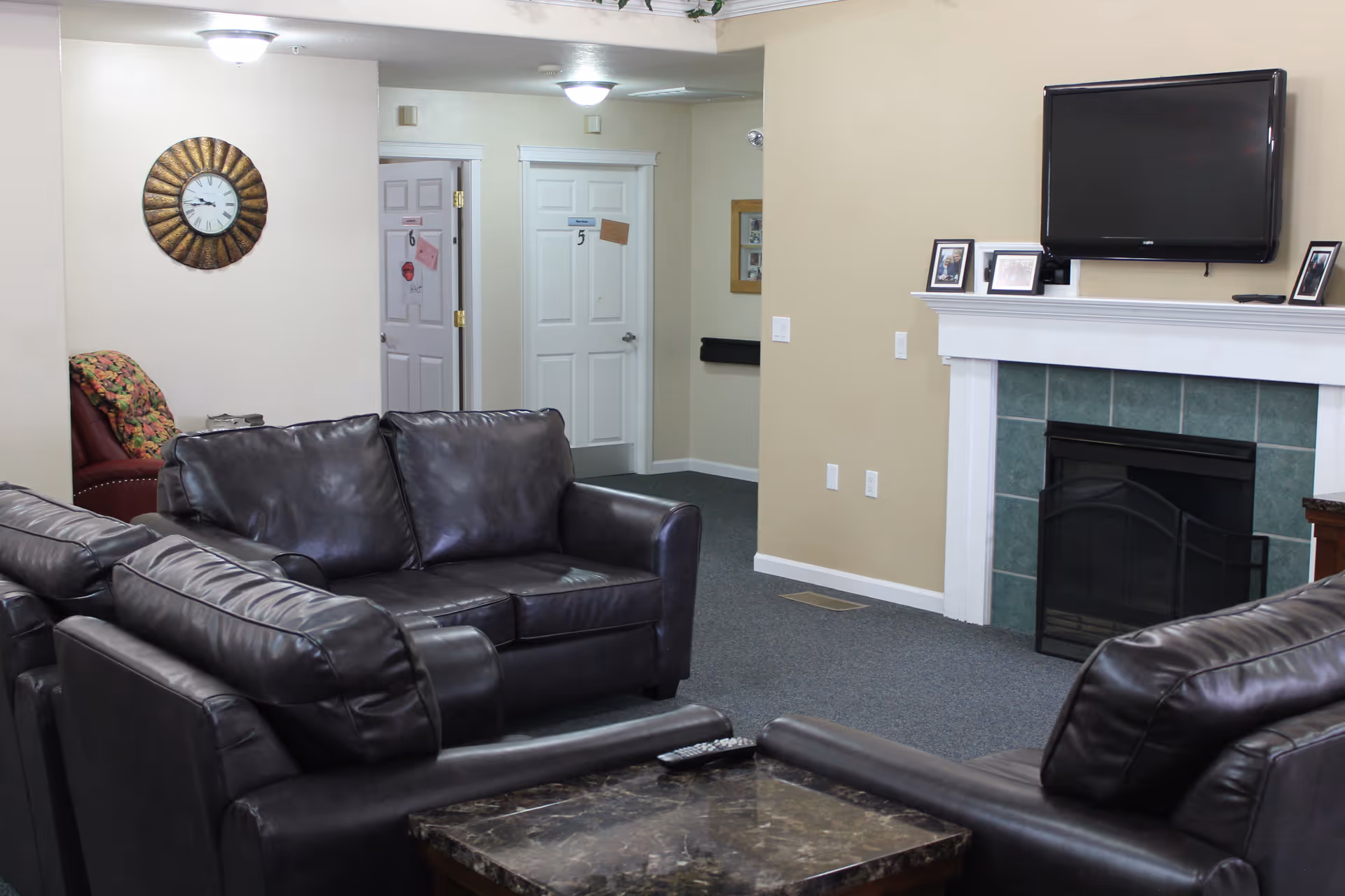 A cozy living room area with three black leather sofas arranged around a dark marble coffee table. A flat-screen TV is mounted above a white fireplace with green tiles. There are framed photos on the mantel. The walls are beige, and there is a decorative wall clock on one wall. Two white doors with numbers 5 and 6 are visible in the background, along with a red armchair with a colorful blanket draped over it.