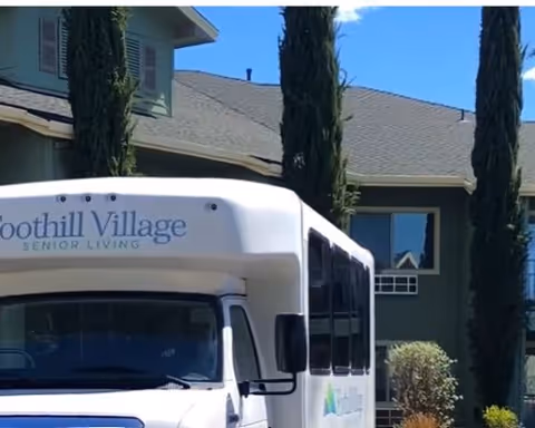 A white shuttle bus with the text 'Foothill Village Senior Living' on the front, parked in front of a green residential building with tall cypress trees and a clear blue sky.
