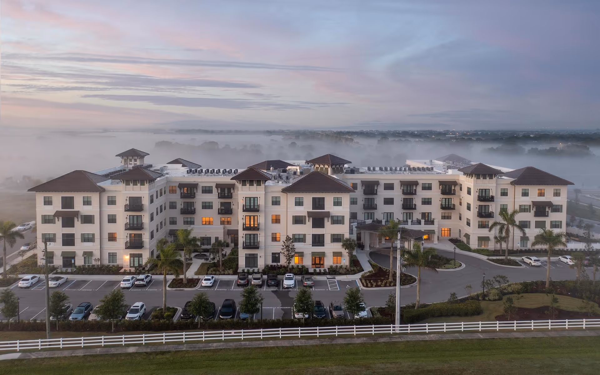 Aerial view of a four-story senior living building with balconies, a parking lot, landscaped palms, and morning fog.