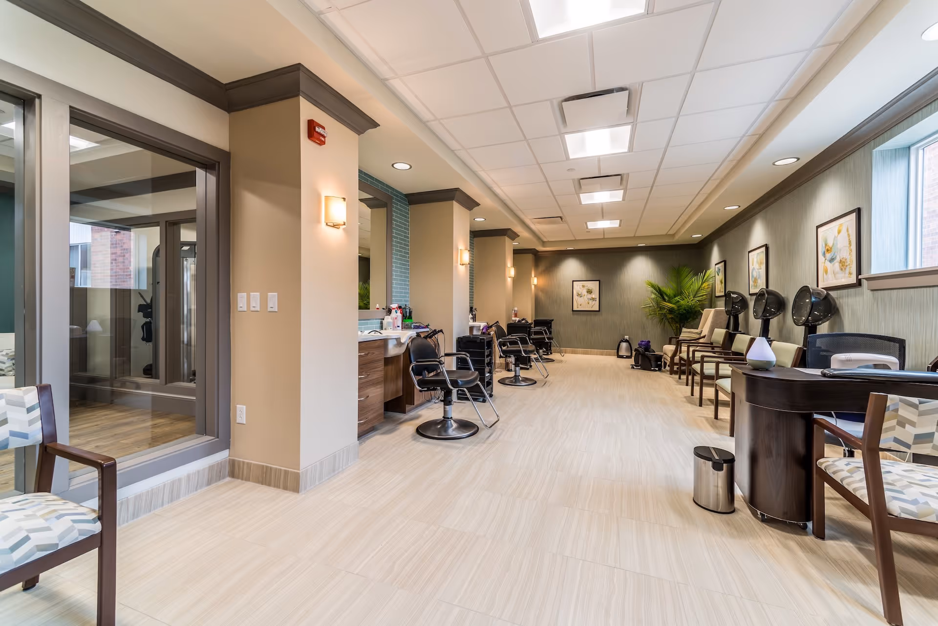 Interior view of a senior living facility's salon area with multiple styling chairs, hair dryers, and a reception desk. The room has light-colored tiled flooring, beige walls with crown molding, framed floral artwork, and a large window letting in natural light. There is a glass partition on the left side showing an adjacent room with exercise equipment.