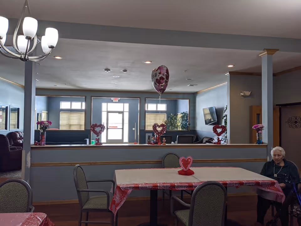 Interior view of a common area in a senior living facility decorated with Valentine's Day themed items including heart-shaped decorations and a balloon. There are tables covered with red and white tablecloths, chairs around the tables, and an elderly woman sitting on the right side. The room has blue walls, a chandelier, and a TV mounted on the wall in the background.