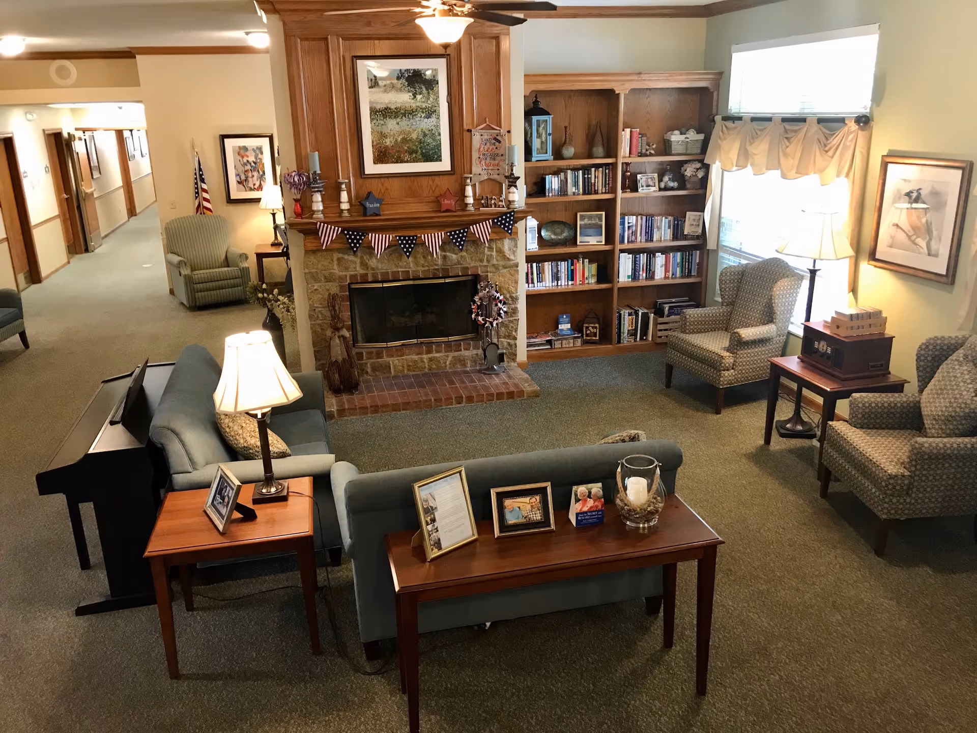 A cozy living room area in a senior living facility with a stone fireplace decorated with patriotic bunting. There are several armchairs and sofas arranged around the fireplace, a wooden bookshelf filled with books, framed pictures on tables, a piano, and lamps providing warm lighting. The room has carpeted floors and light-colored walls with windows letting in natural light.
