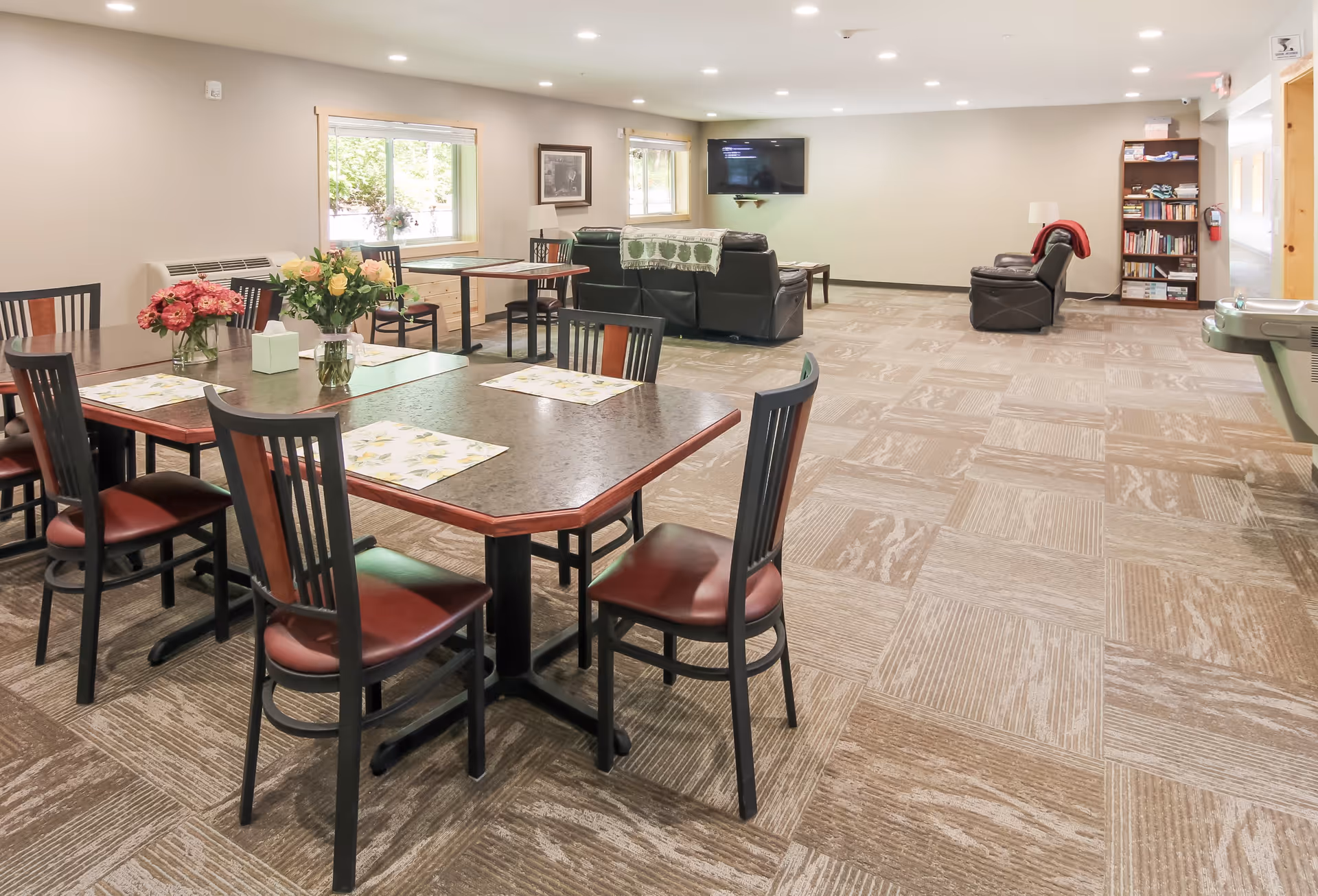 A spacious common area in South Torch Assisted Living featuring multiple tables with chairs, floral centerpieces, a seating area with leather couches and armchairs, a wall-mounted TV, a bookshelf, and large windows letting in natural light.