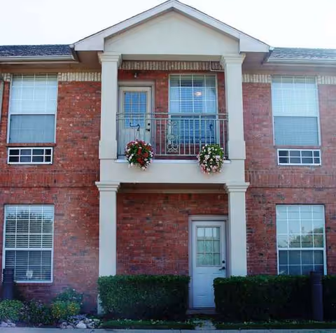 Brick senior living building front with central balcony and flower baskets.