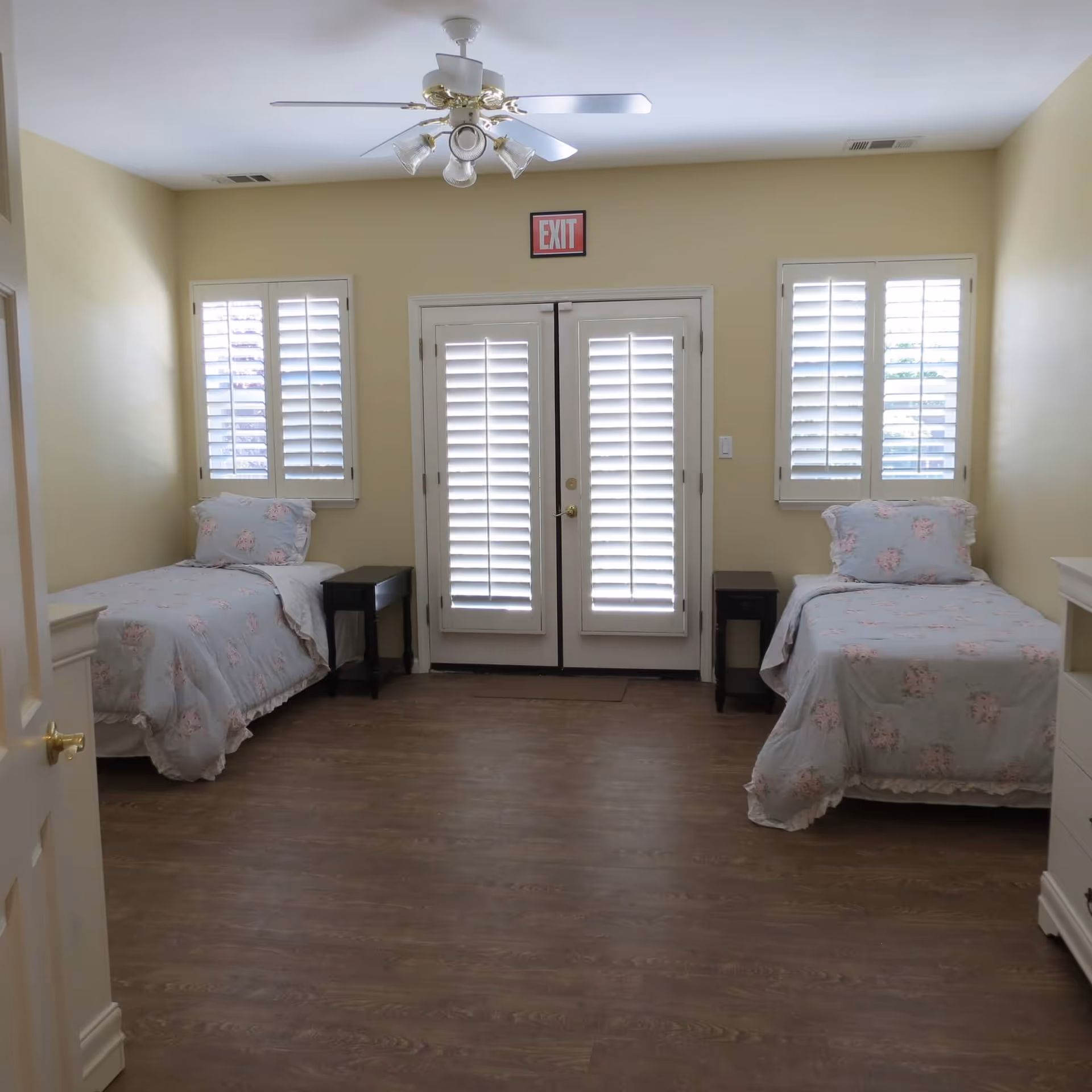 Bright shared bedroom with two twin beds, nightstands, a ceiling fan, and shuttered French doors with an EXIT sign above.