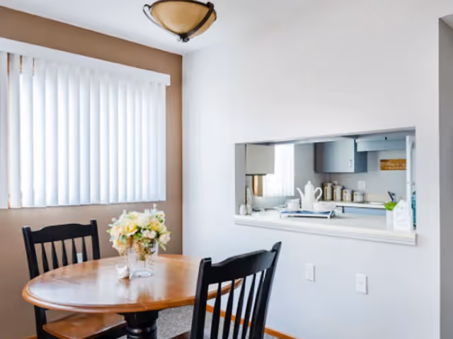 A small dining area with a round wooden table and two black chairs. A vase with light-colored flowers is placed on the table. There is a window with vertical blinds on the left wall, and a pass-through window opens into a kitchen area with white cabinetry and various kitchen items visible.