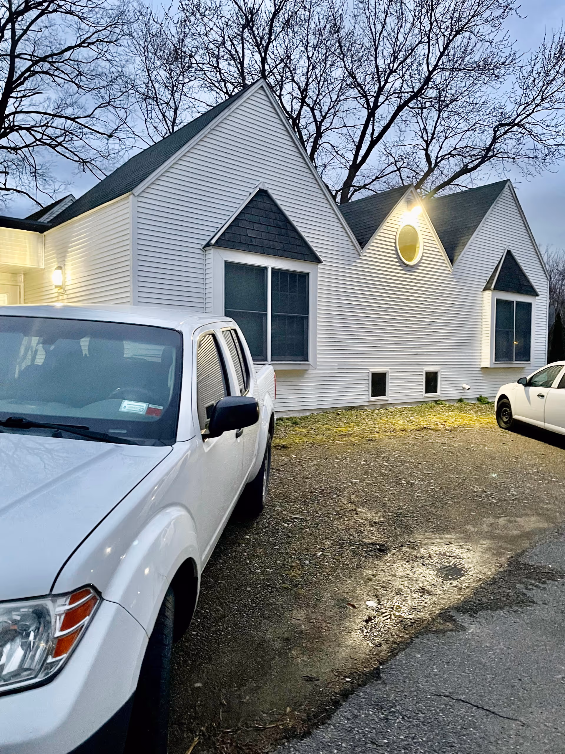 White single-story building with steep gables and exterior lights, viewed from a gravel parking area with two white vehicles in front at dusk.