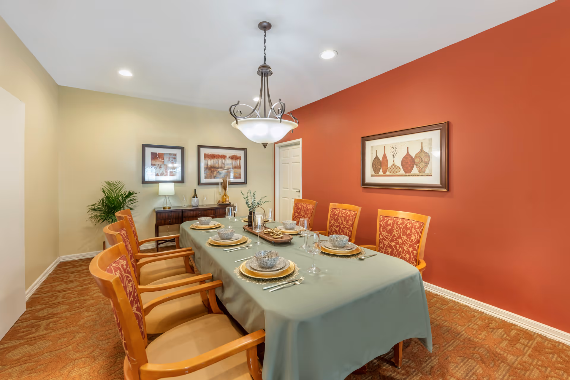 A dining room with a long table covered with a green tablecloth set for six people with plates, bowls, glasses, and silverware. The room has six wooden chairs with red patterned cushions. The walls are painted beige and red, with framed artwork hanging on them. There is a sideboard with a lamp, decorative items, and a potted plant in the corner. A chandelier hangs above the table.