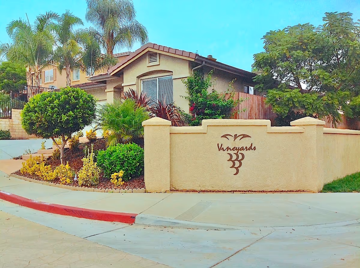 Entrance wall and landscaped yard in front of a single-story stucco home with a 'Vineyards' sign and grape logo.