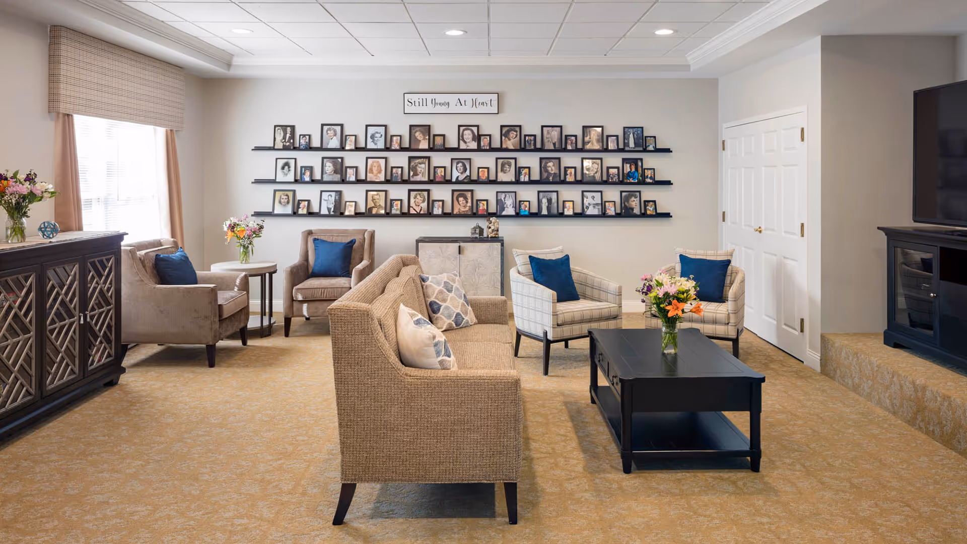 A cozy living room with beige carpet and neutral-toned furniture including a sofa, armchairs with blue cushions, and a black coffee table with a flower vase. The wall features three shelves filled with framed photos and a sign above that reads 'Still Young At Heart'. There is a large window with curtains on the left and a TV on a black stand on the right.
