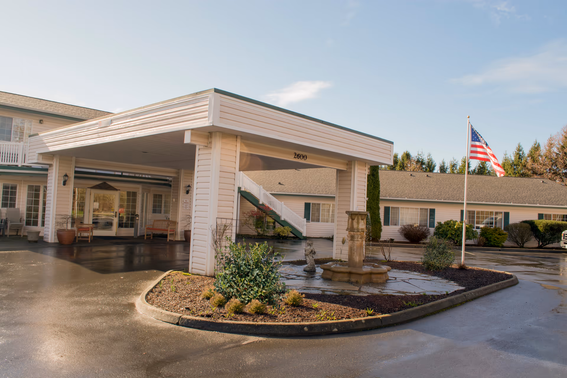 Exterior view of Brookdale Fairhaven senior living facility entrance with a covered drop-off area, a small landscaped island with a fountain and statue, and an American flag on a flagpole. The building is light-colored with green shutters and a two-story section visible in the background.