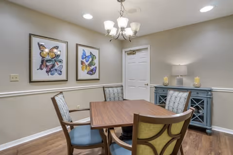 A small dining room with a wooden table and four chairs with patterned upholstery. The room has light beige walls with white trim, two framed butterfly artworks on one wall, a blue sideboard with a lamp and two candles, and a white door. A chandelier with five lights hangs from the ceiling.