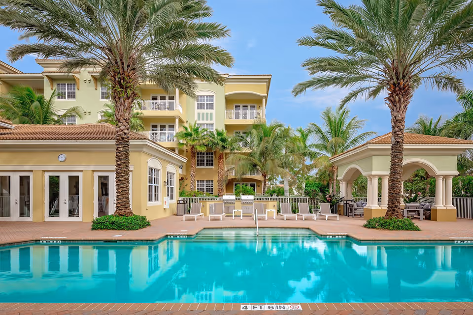 Outdoor swimming pool area at Riverwalk Pointe with clear blue water, surrounded by palm trees and lounge chairs. There is a building with large windows and a clock on the wall to the left, and a covered seating area with columns to the right. The sky is clear and blue.