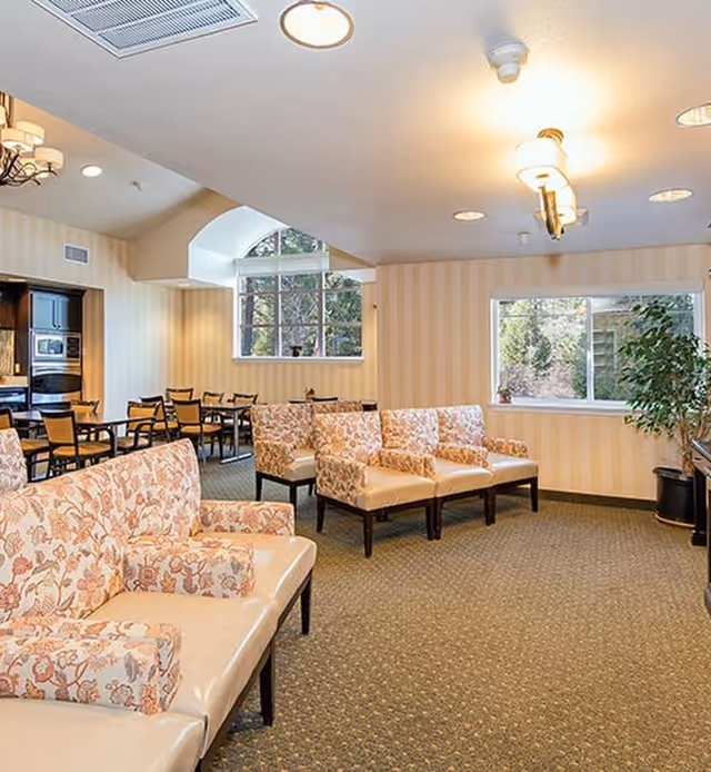 A well-lit common area with floral patterned cushioned chairs and sofas arranged on a carpeted floor. There are large windows letting in natural light, a potted plant near one window, and a dining area with tables and chairs in the background. The walls have a light striped wallpaper and ceiling lights provide additional illumination.